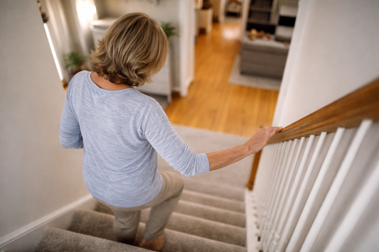 Person in their 50s or 60s walking down a staircase at home while holding the railing, viewed from behind with natural lighting and a realistic everyday setting