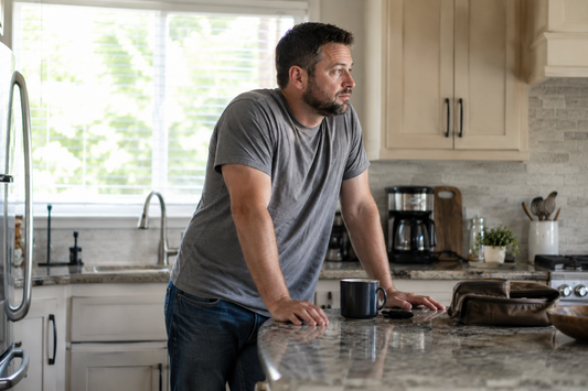 Middle-aged man standing in a kitchen with coffee in the morning, representing lingering fatigue, stiffness, and not feeling fully recovered after rest