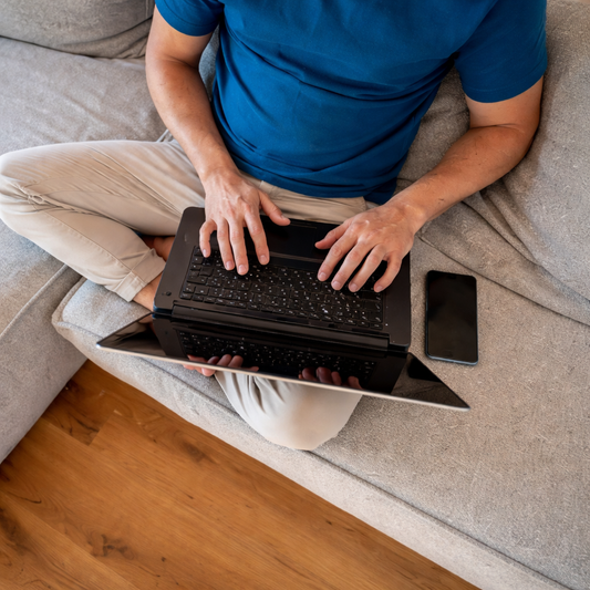 Man sitting cross-legged on a couch using a laptop with a phone nearby, casual everyday home setting with natural lighting