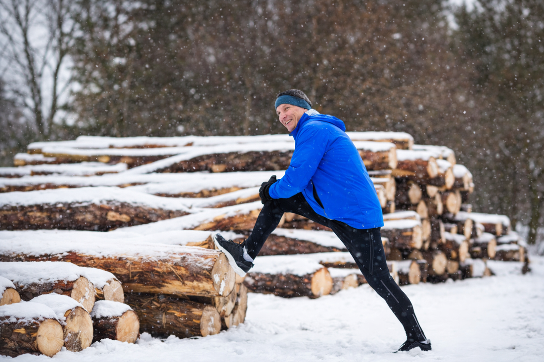 man stretching leg on stacked logs outdoors in snowy winter landscape before walking