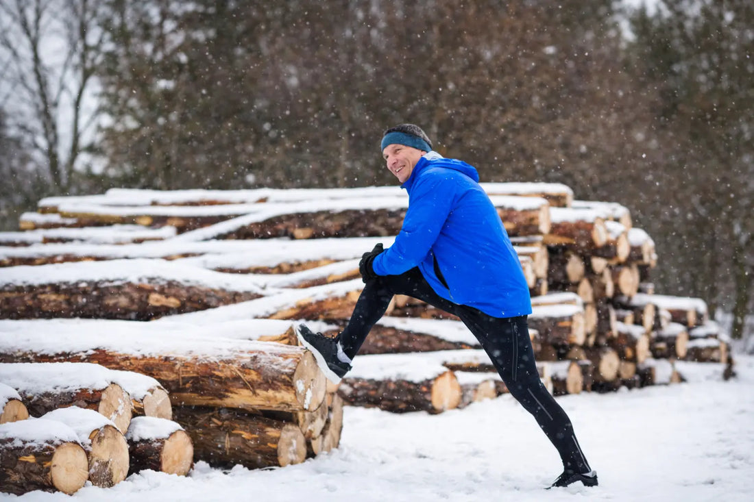 man stretching leg on stacked logs outdoors in snowy winter landscape before walking