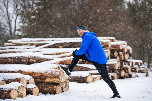 man stretching leg on stacked logs outdoors in snowy winter landscape before walking