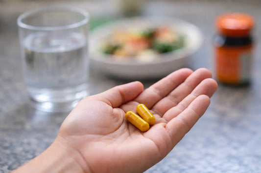 Hand holding two turmeric supplement capsules in a kitchen with a glass of water and a meal blurred in the background.