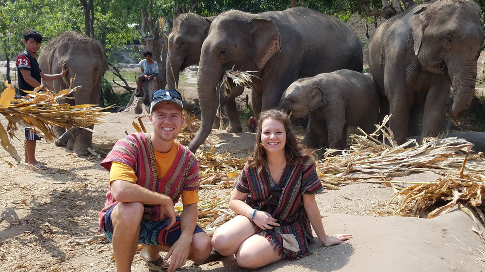 A man and a woman sitting in front of a group of elephants at a sanctuary or farm, with people and dried palm leaves in the background.