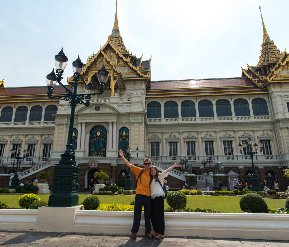 Two people posing in front of the Grand Palace in Bangkok, Thailand, with their arms raised in excitement. The iconic golden-roofed palace building dominates the background under a bright sky, with green lawns and decorative lamp posts in the foreground.