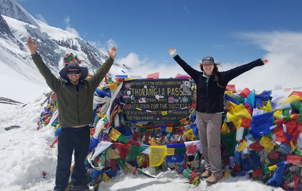 Two people standing in snow with arms raised in front of a mountain pass sign and colorful prayer flags, with snowy peaks and blue sky in the background.