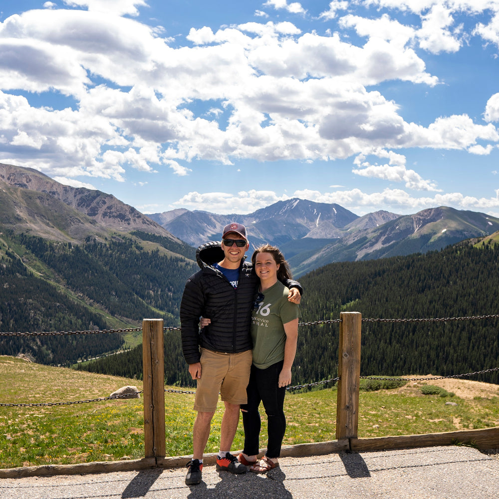 A couple posing together outdoors on a mountain trail overlook with a vast blue sky, white clouds, and green and gray mountain ranges in the background.