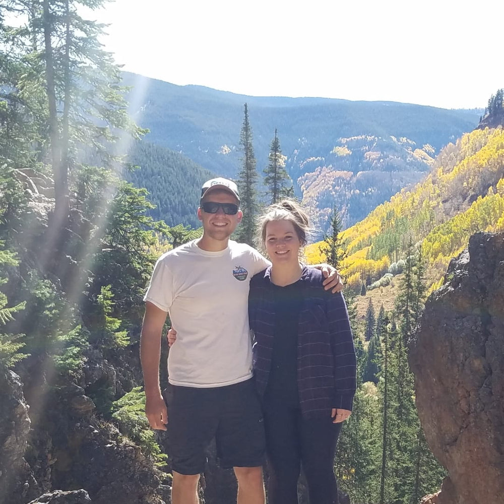 A man and a woman standing together on large rocks in a mountainous outdoor setting with trees and autumn-colored hills in the background.