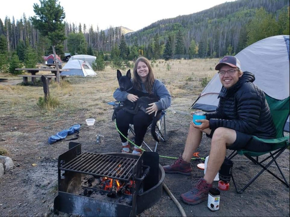 A family camping scene with two people sitting in folding chairs by a campfire, a dog in the center, a white tent behind them, and a forested mountain in the background.