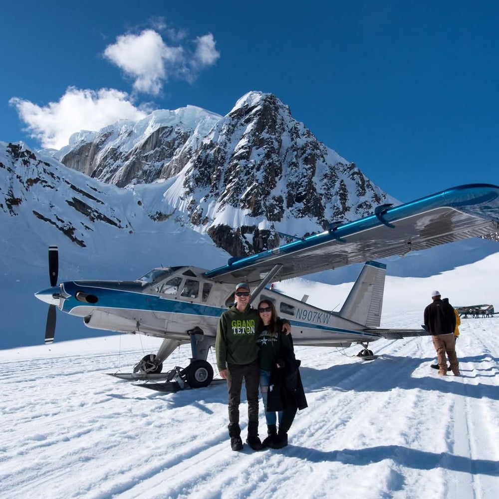 A small blue and white seaplane parked on a snowy mountain pass with snow-covered peaks and a deep blue sky in the background. Two people stand in the foreground near the plane, and another person is visible further back on the right.